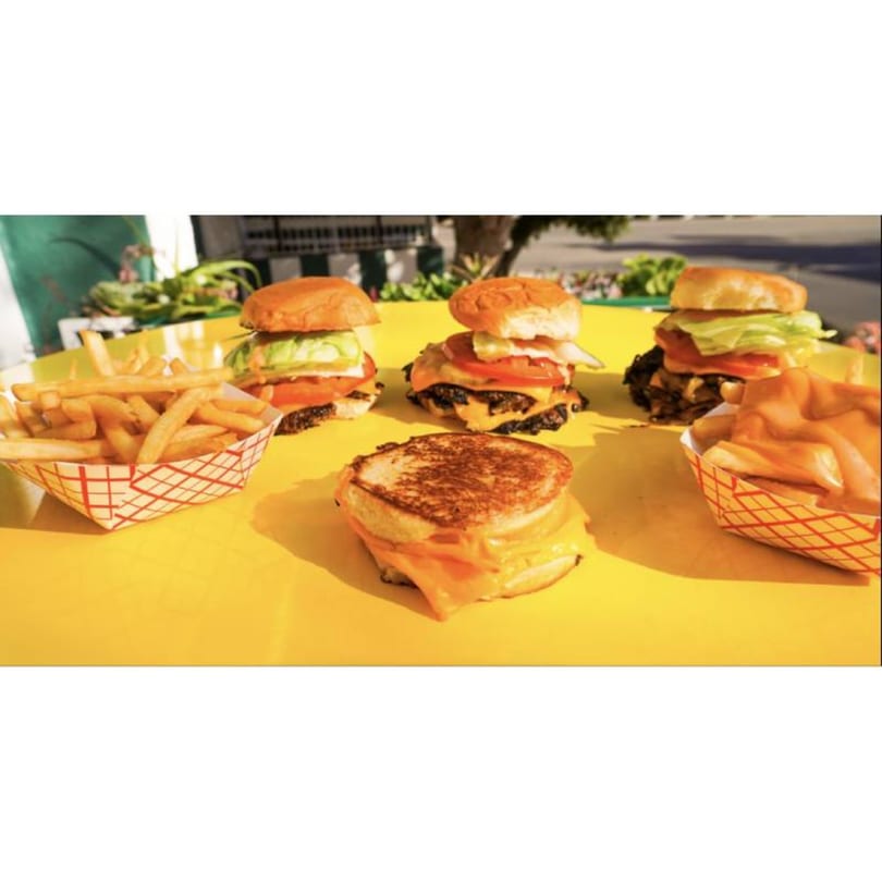 Three Patty Smash burgers, a grilled cheese, and two baskets of fries on a yellow table.