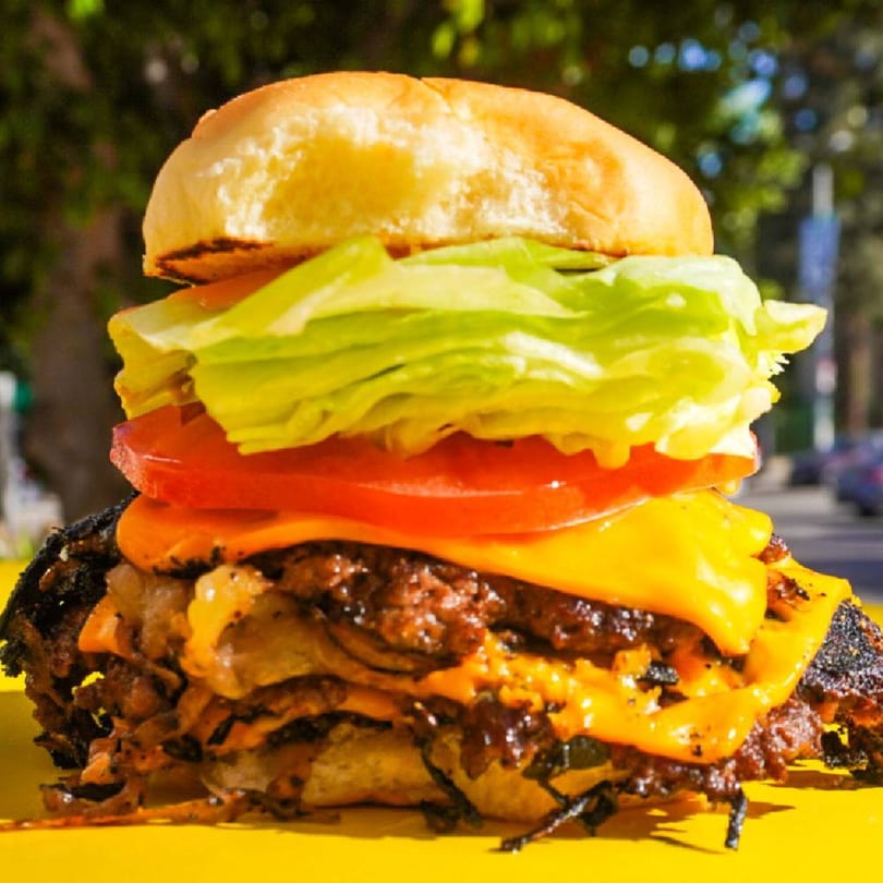 A Patty Smash Brand burger with two patties, melted cheese, lettuce, and tomato on a yellow surface.